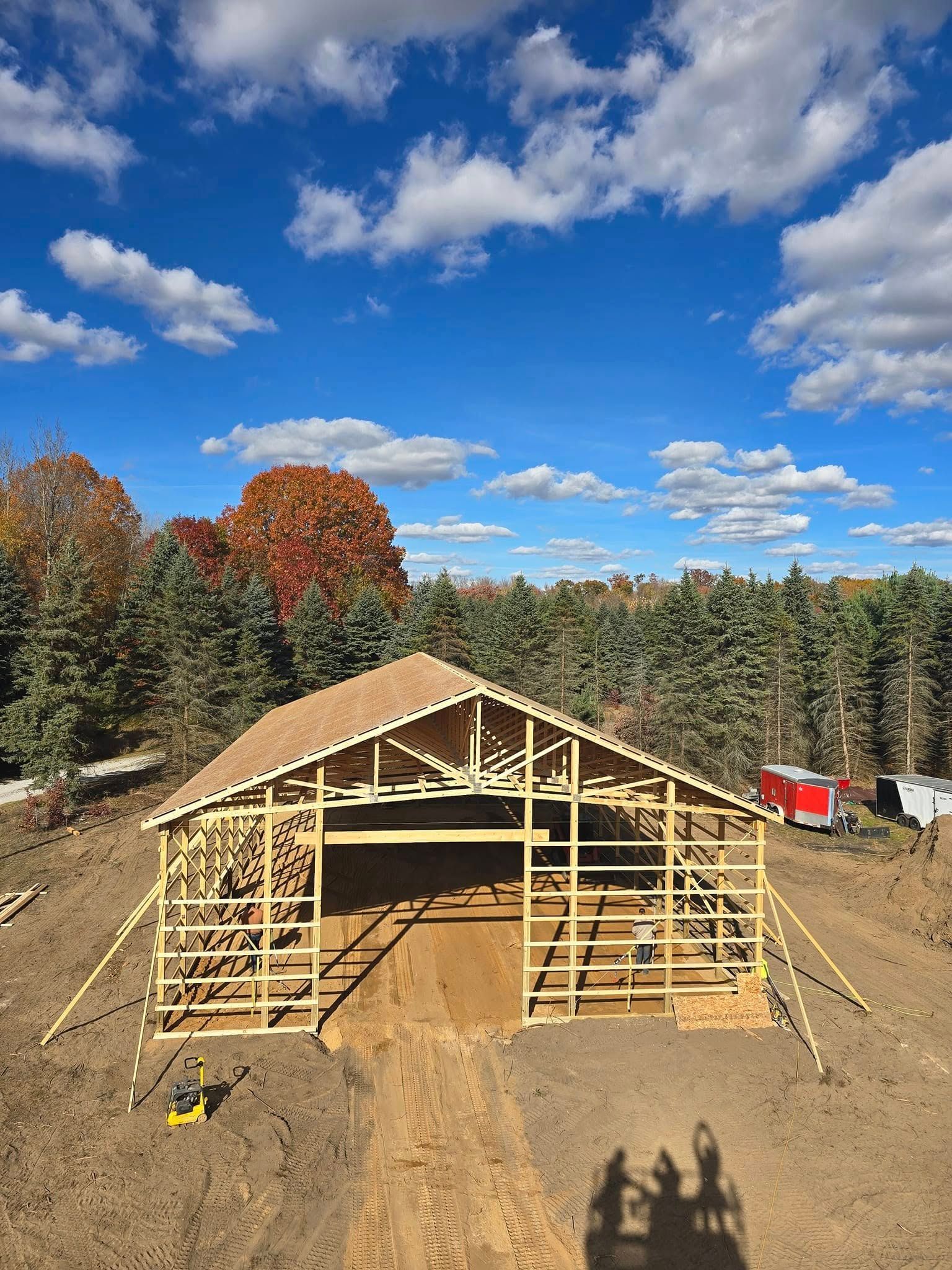 Wooden barn frame under construction with a blue sky and trees in the background.