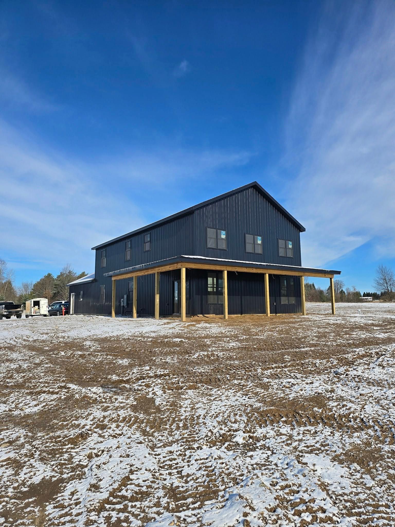 Two-story black building under construction with a wraparound porch, set in a snowy field under a blue sky.