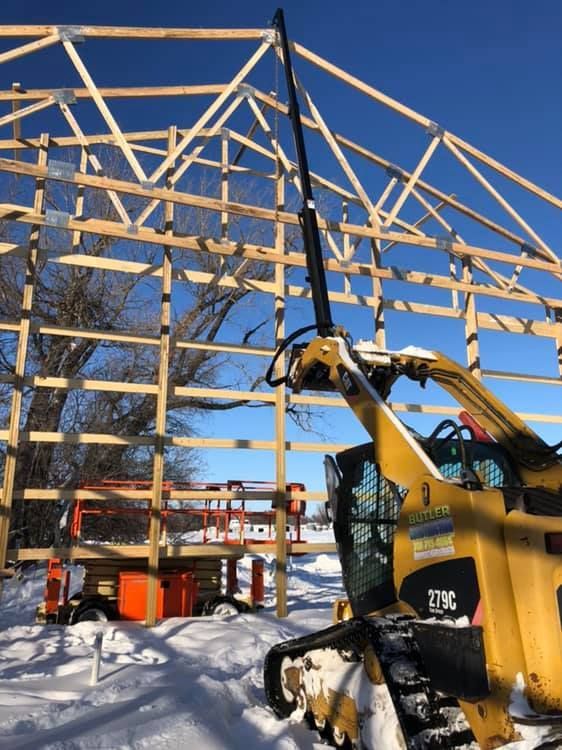 A tracked skid steer with an extended lift arm at a wood frame barn under construction in a snowy setting.