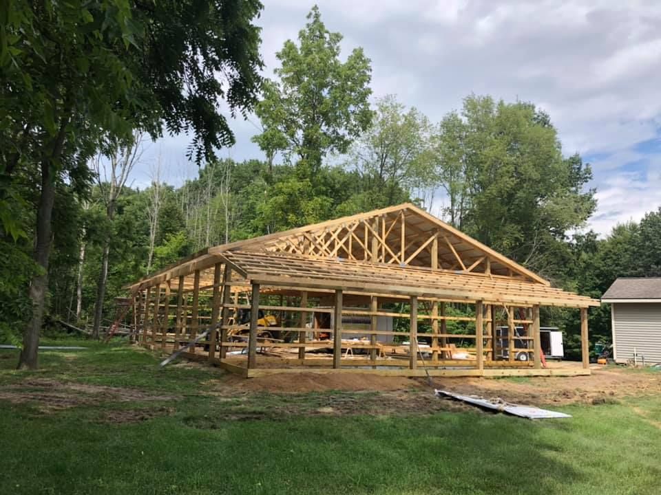 Wooden frame of a building under construction, set in a grassy yard with trees in the background.