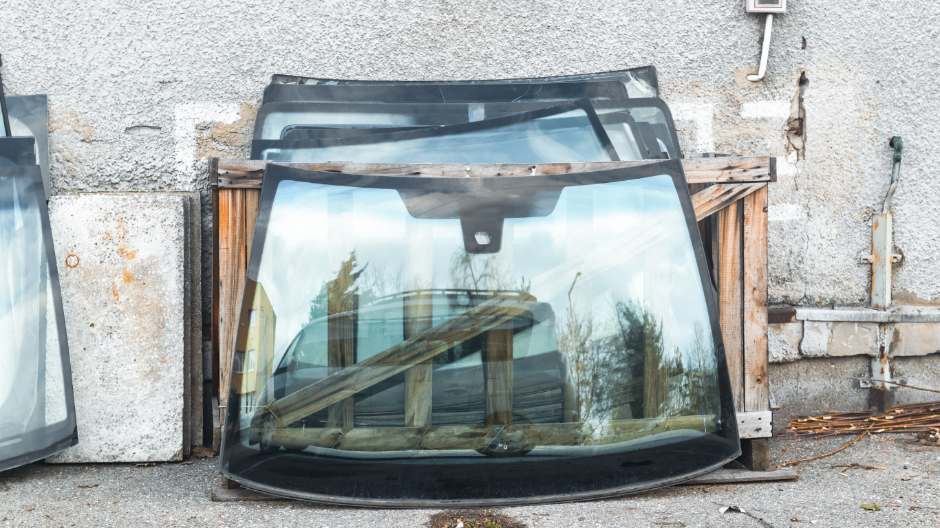 Windshield glass sheets stacked against a weathered wall, reflecting trees and sky.