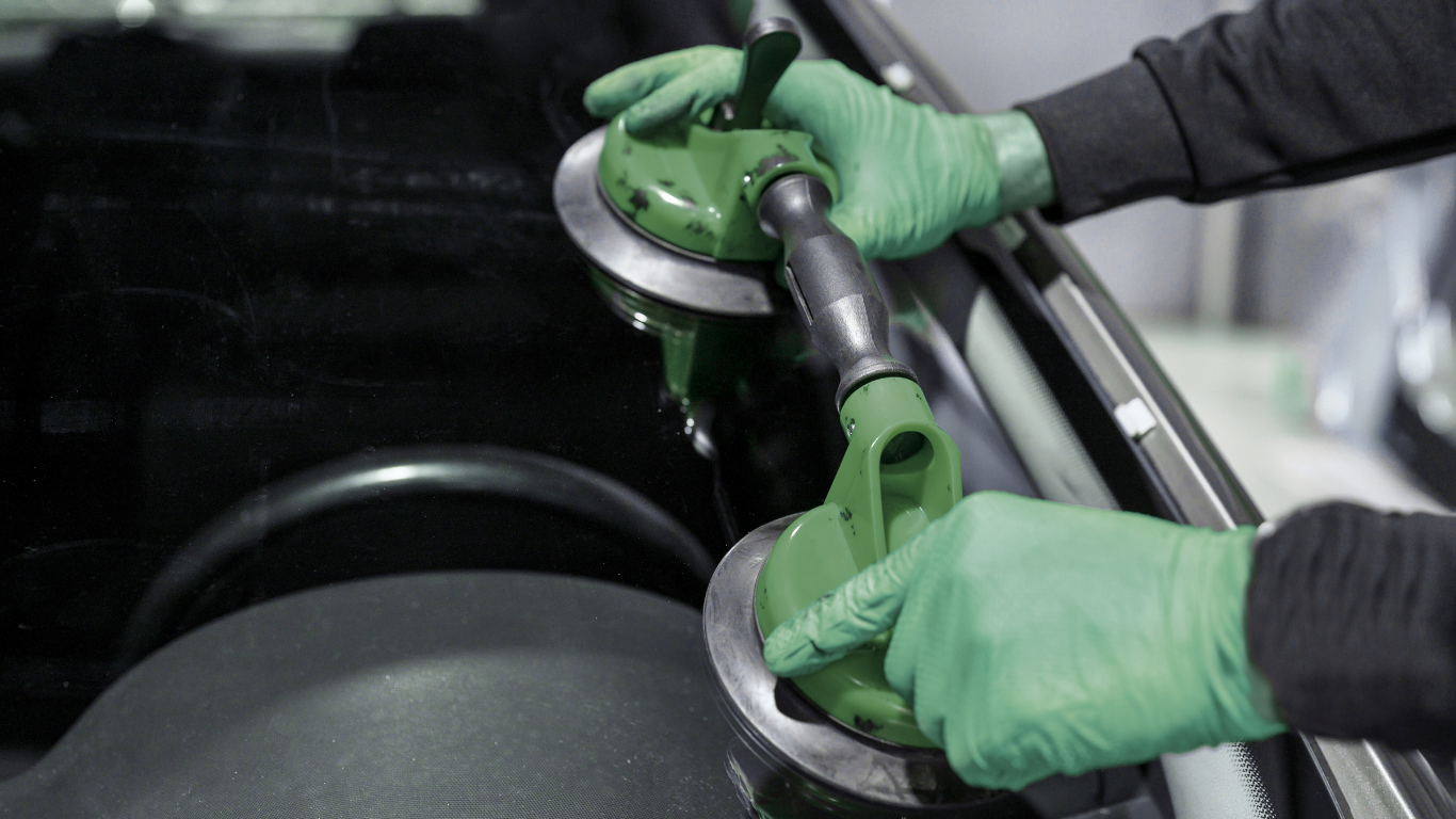 Green-gloved hands using suction cups to remove a car windshield in a repair shop.