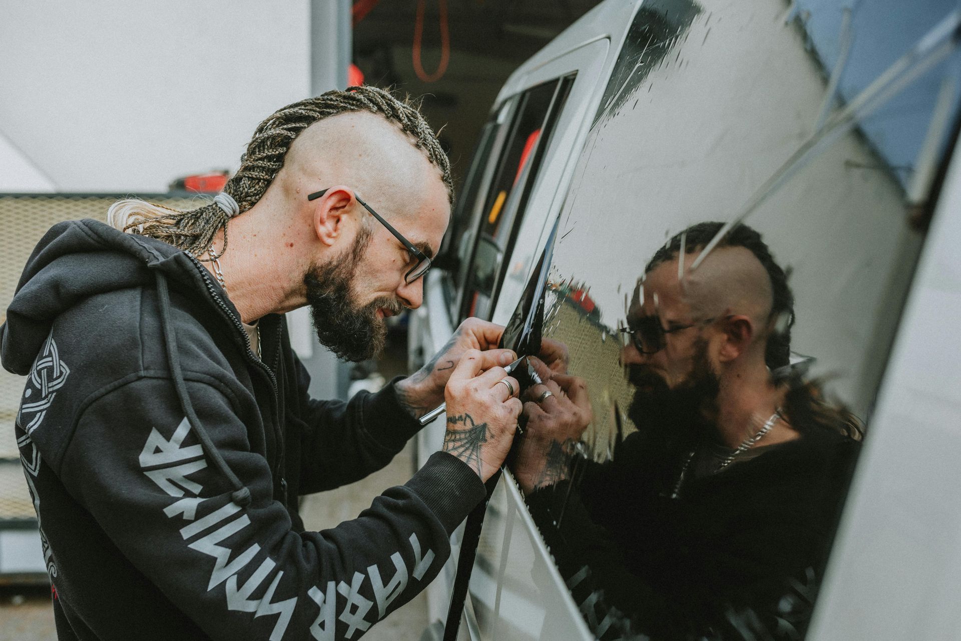 Person with mohawk and beard working on a vehicle window, reflection visible.