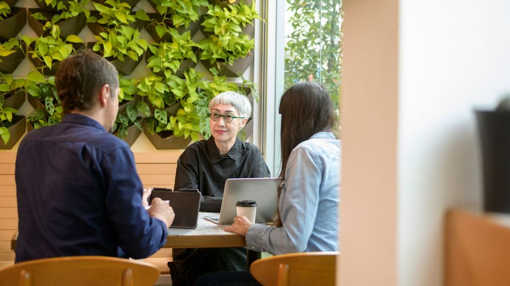 Three people seated at a table in a cafe, discussing. Green wall in background.