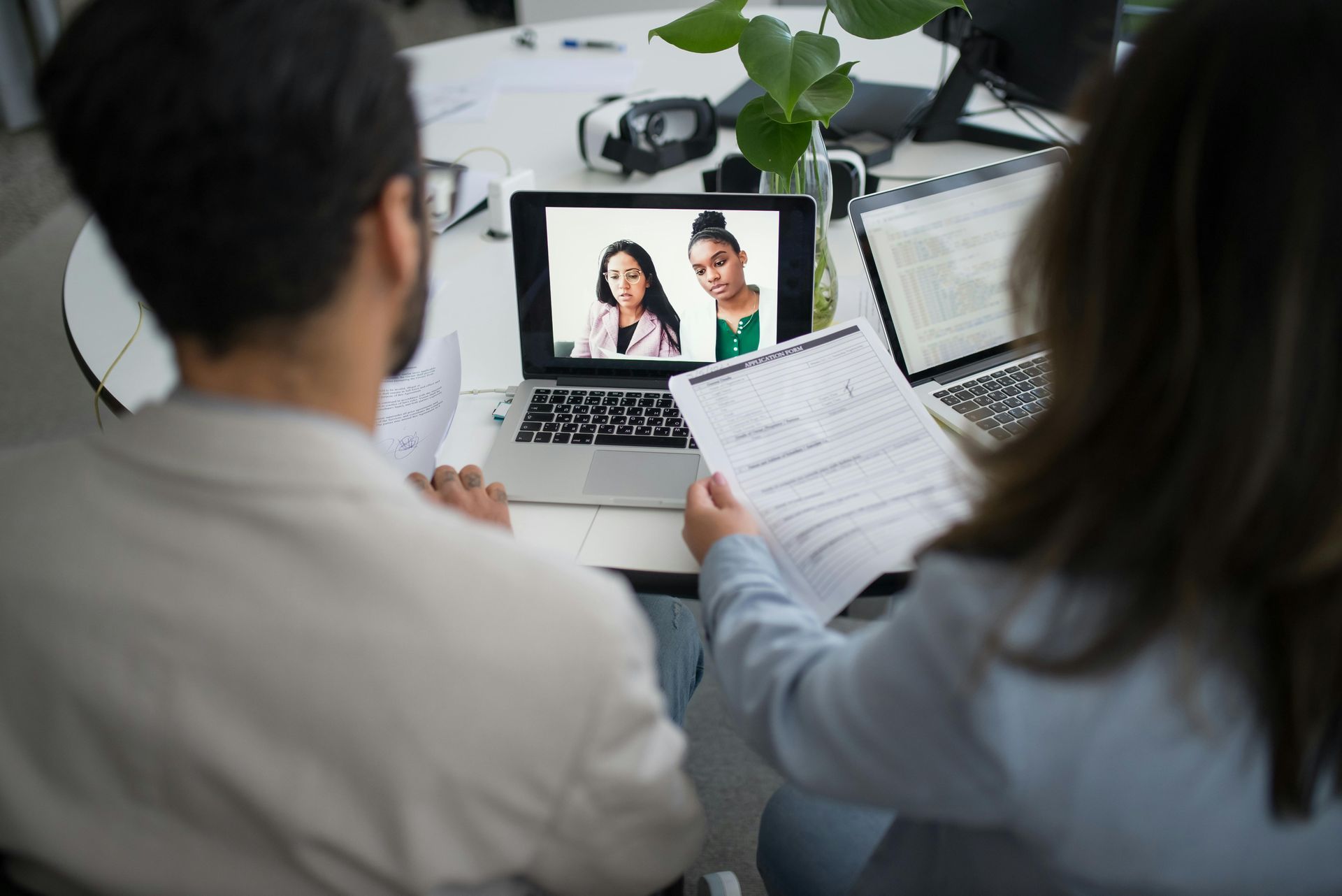Two people at a desk in a business setting having a video conference with two women on a laptop screen.