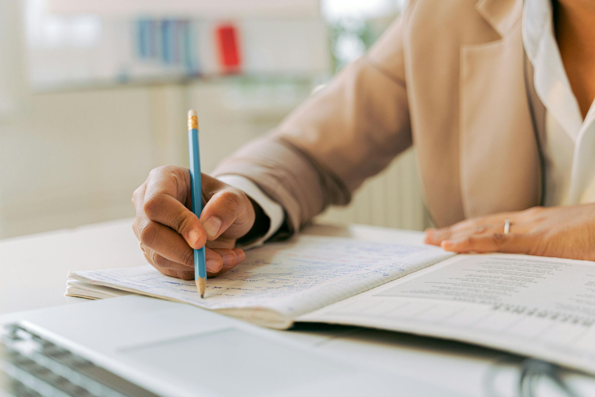 Person writing with a pencil in a notebook at a desk, wearing a light brown blazer.