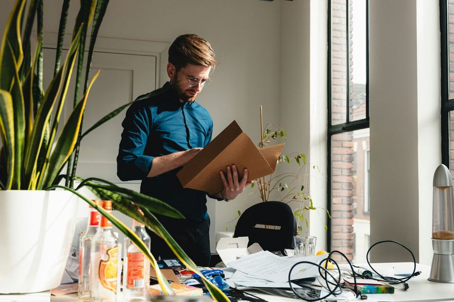 Man in glasses, looking at papers in an office. A large plant and a window are behind him.