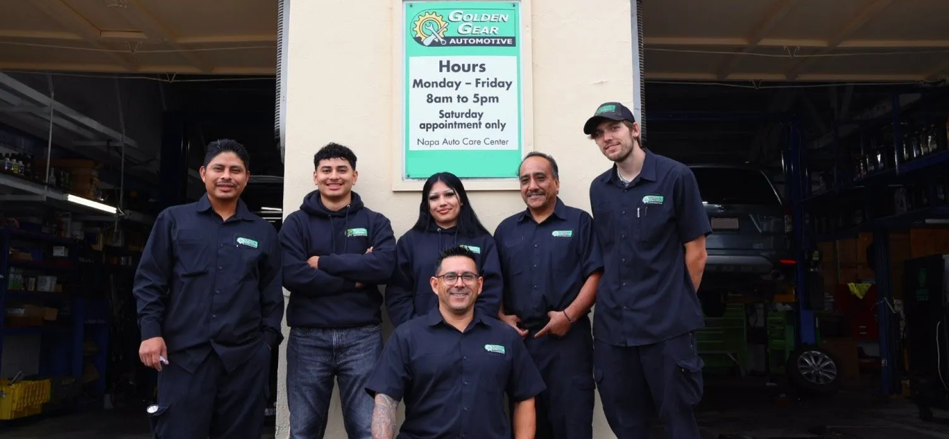 Group of six people smiling in front of a building with a sign. The people wear work uniforms. | Golden Gear Automotive