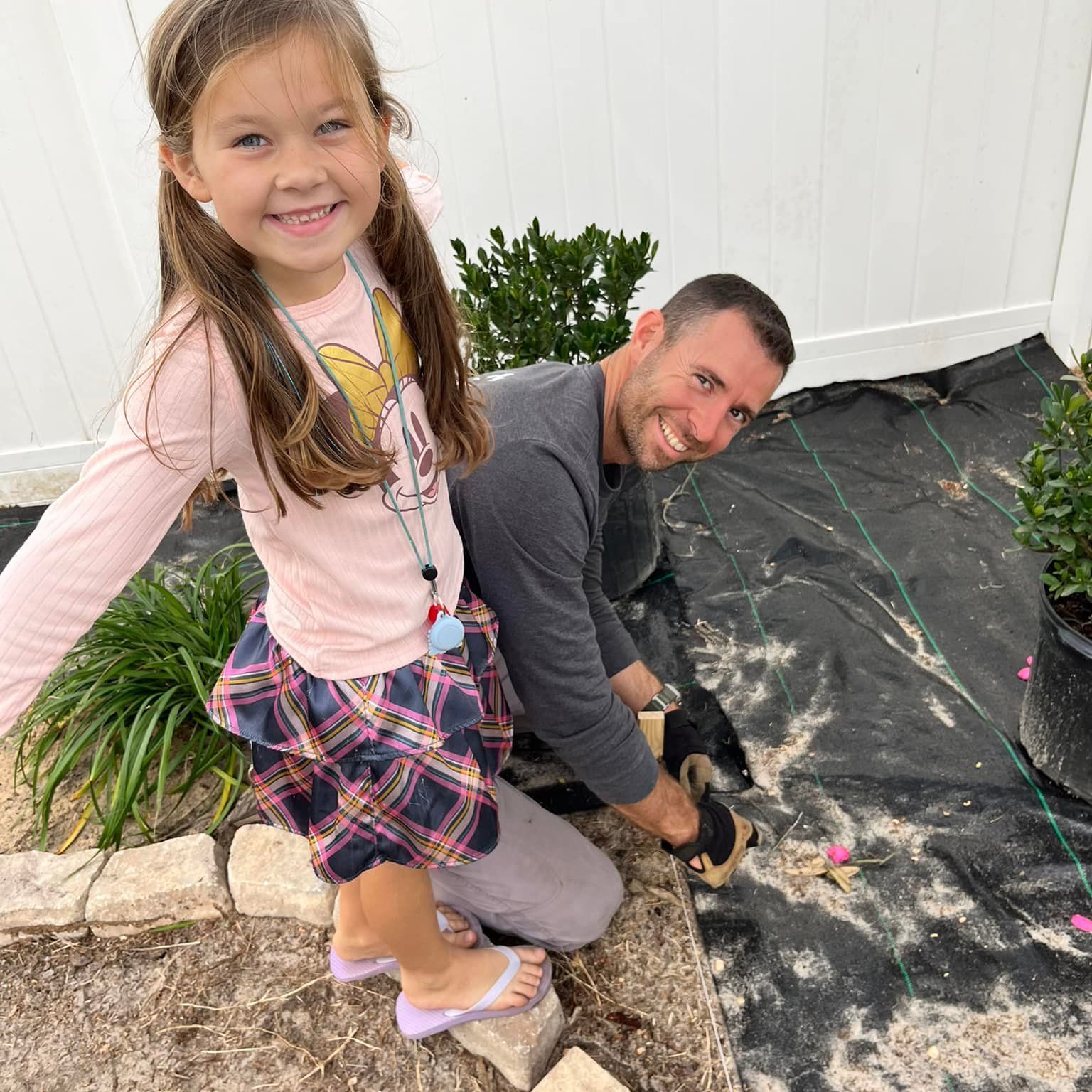 A little girl is standing next to a man kneeling down in the dirt.