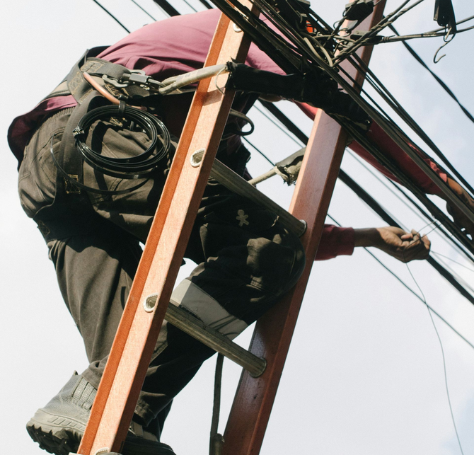 A worker in dark clothing and a safety harness climbing an orange ladder to repair overhead utility lines.