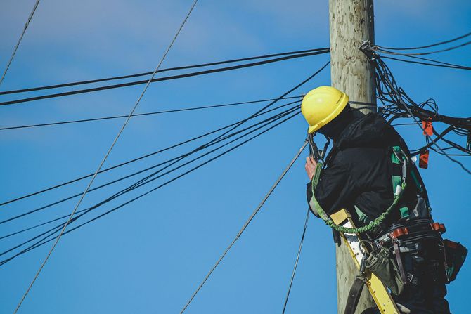 A utility worker in a yellow hard hat and safety harness works on electrical wires atop a utility pole against a blue sky.