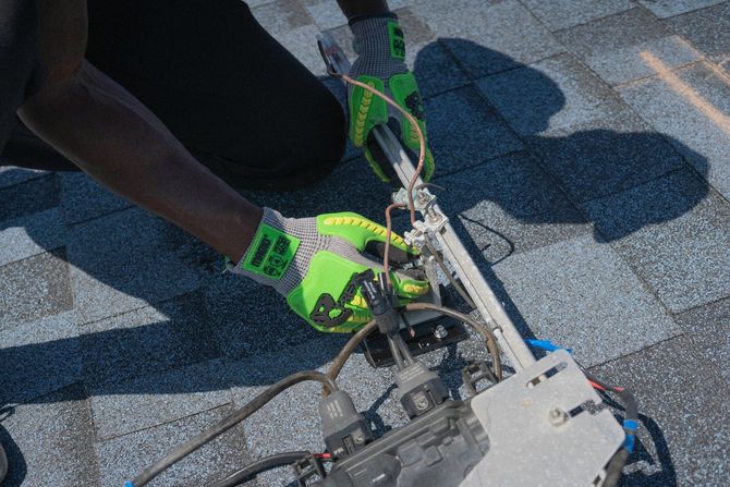 A person wearing high-visibility work gloves secures electrical wiring to a rail system mounted on a shingled roof.