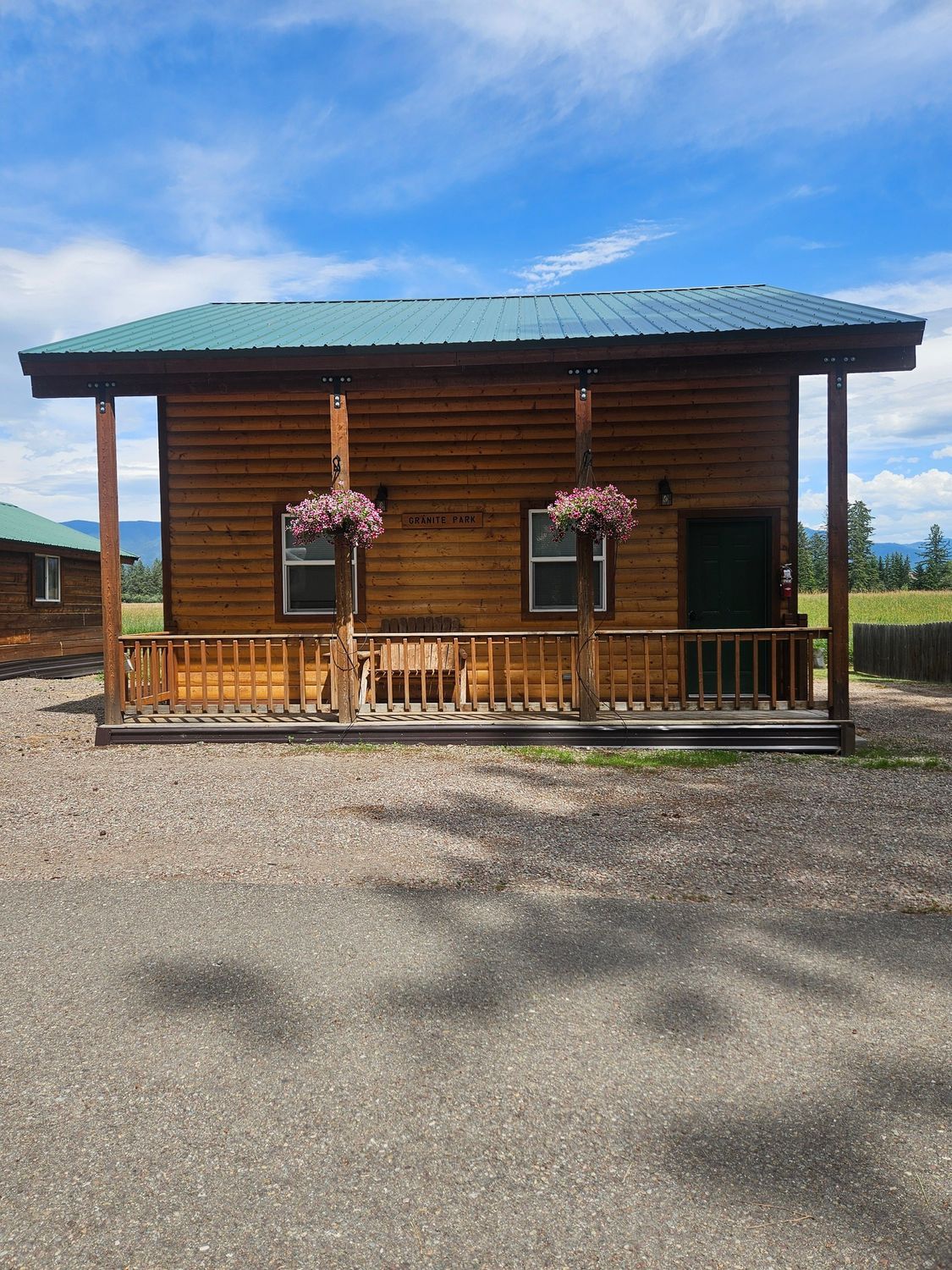 Wooden cabin with porch, flower baskets, and green roof under a blue sky.