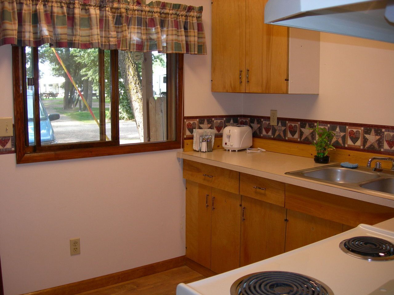 Kitchen with light wood cabinets, window with checkered valance, and white appliances.