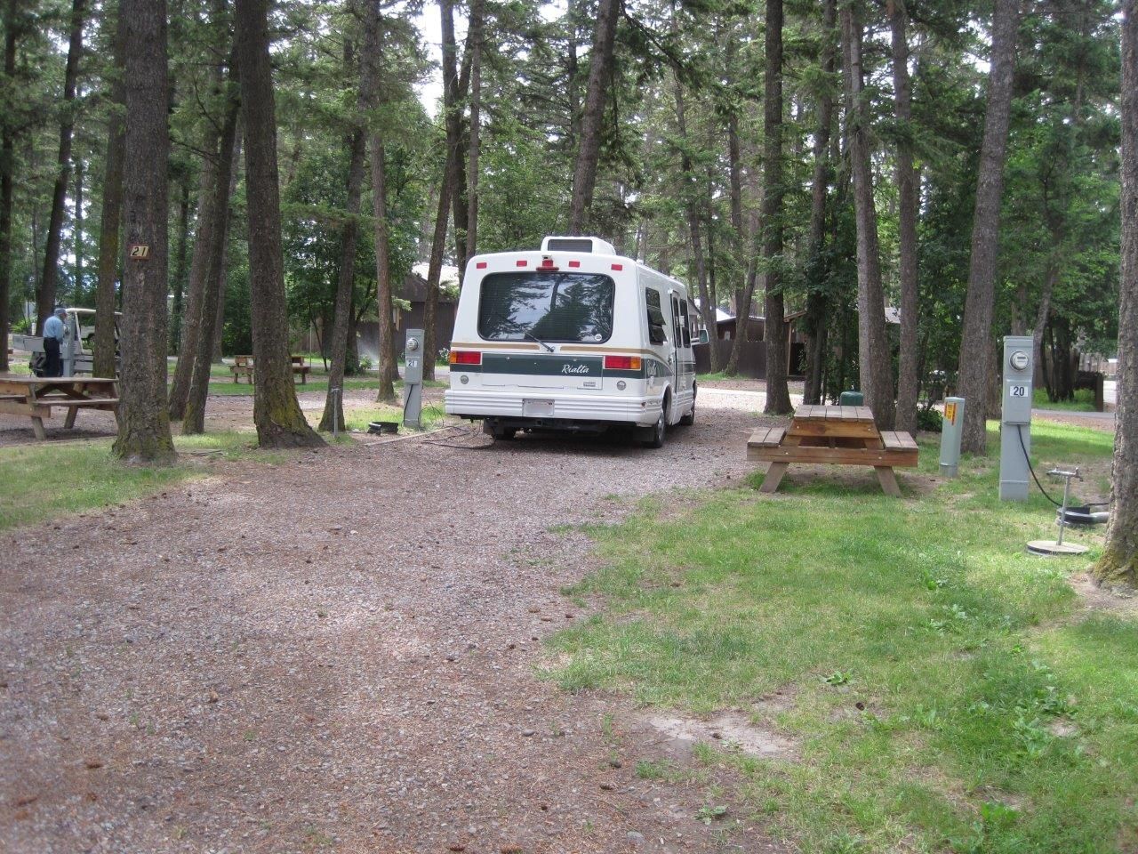 RV parked in a gravel campsite surrounded by trees, with picnic tables and power outlets visible.