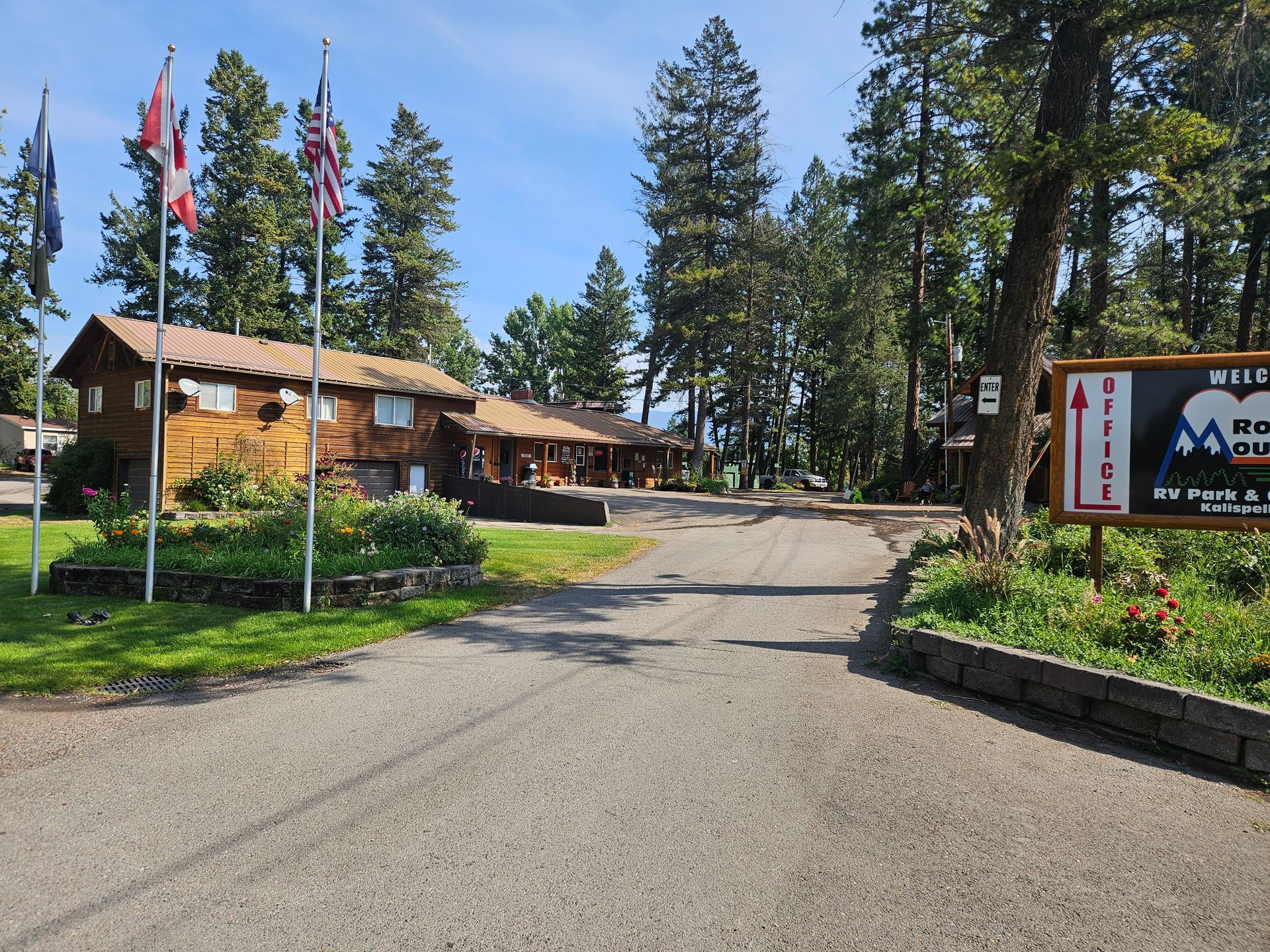 A gravel driveway leads to a log cabin lodge with flags, trees, and a sign that reads 