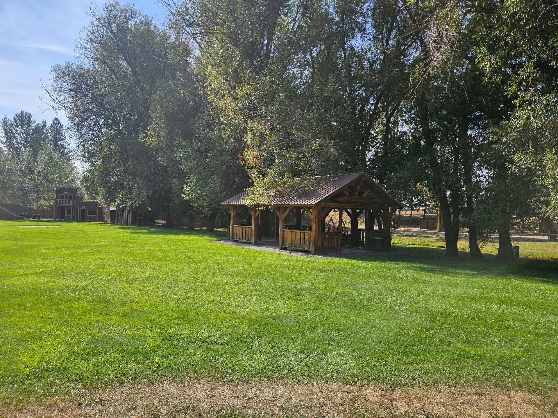 Wooden gazebo on green lawn, surrounded by trees under a blue sky.