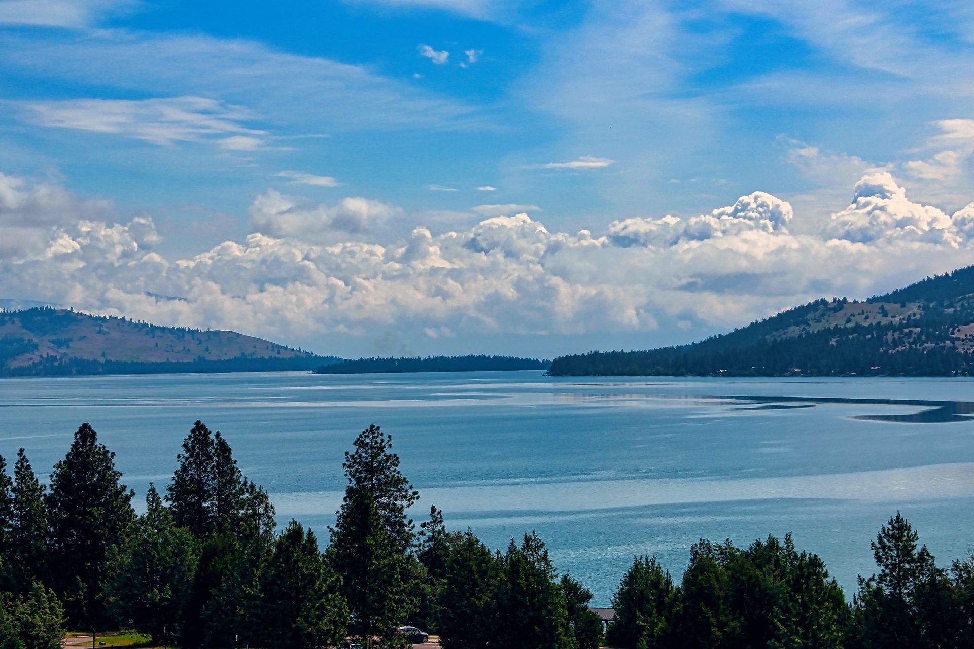 Lake surrounded by tree-covered hills under a blue sky with fluffy white clouds.