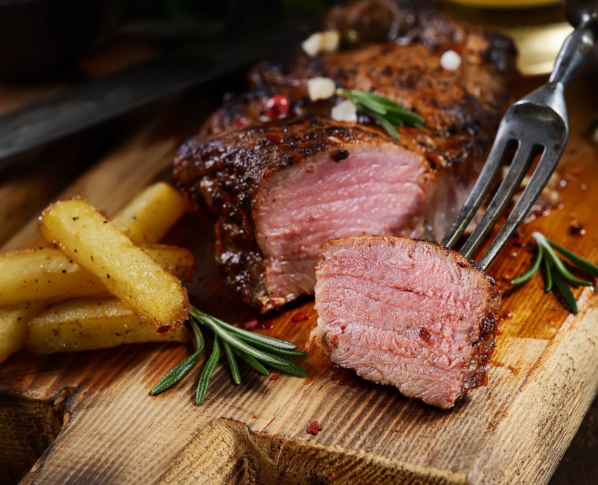 Medium-rare steak and fries on a wooden board with rosemary and a fork.
