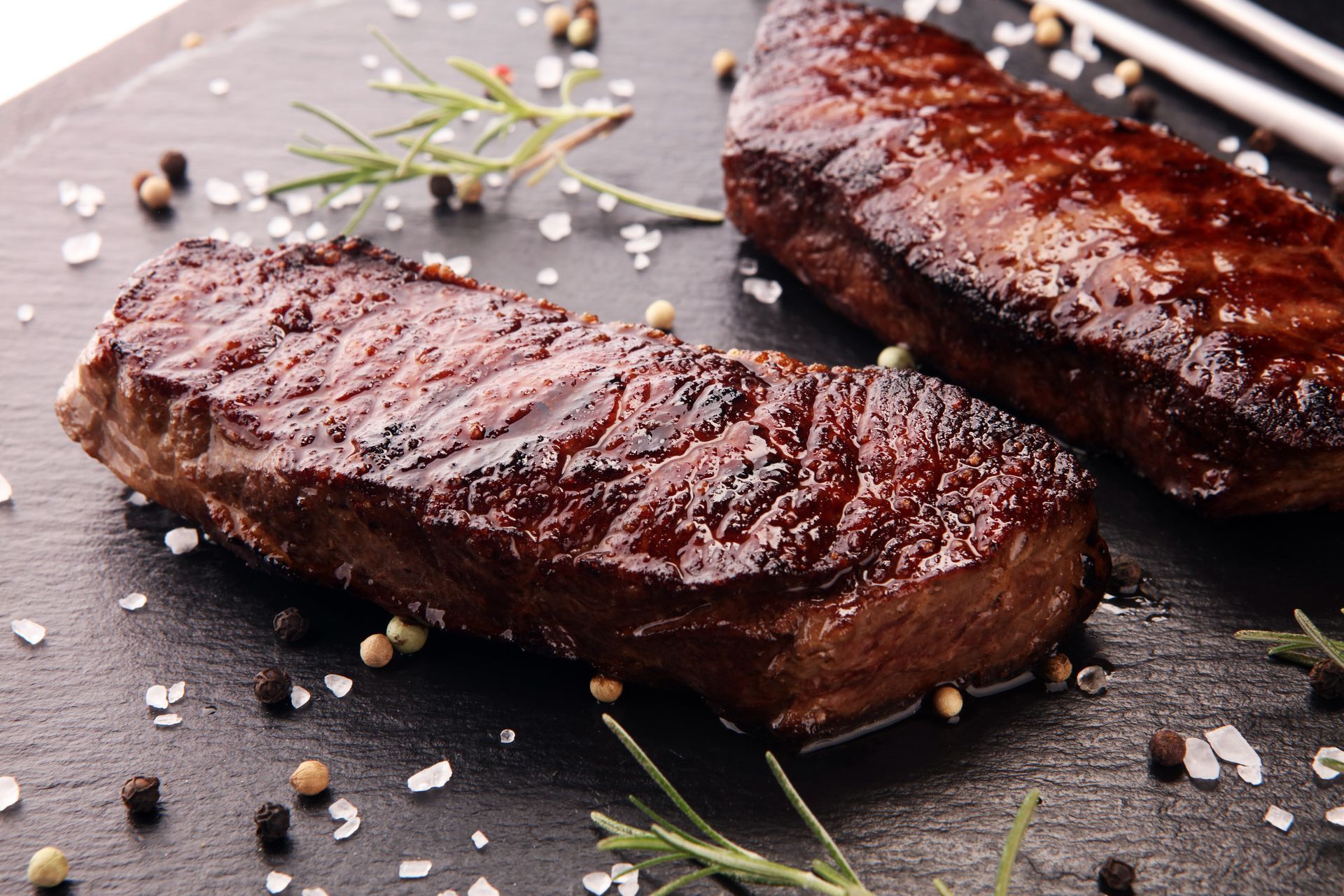 Two grilled steaks on a dark surface, seasoned with salt, pepper, and rosemary.