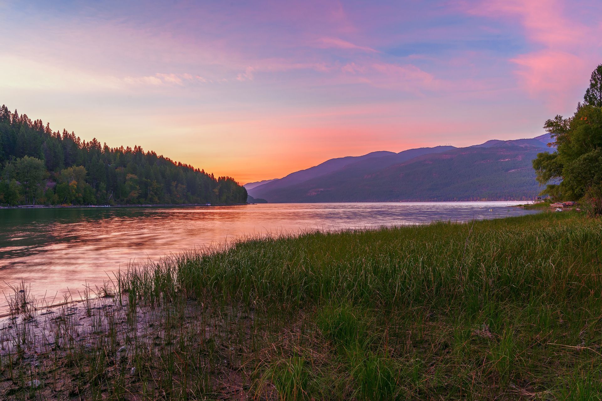 Sunset over lake, pink and purple sky reflects on water. Lush green shoreline, forested hills in distance.