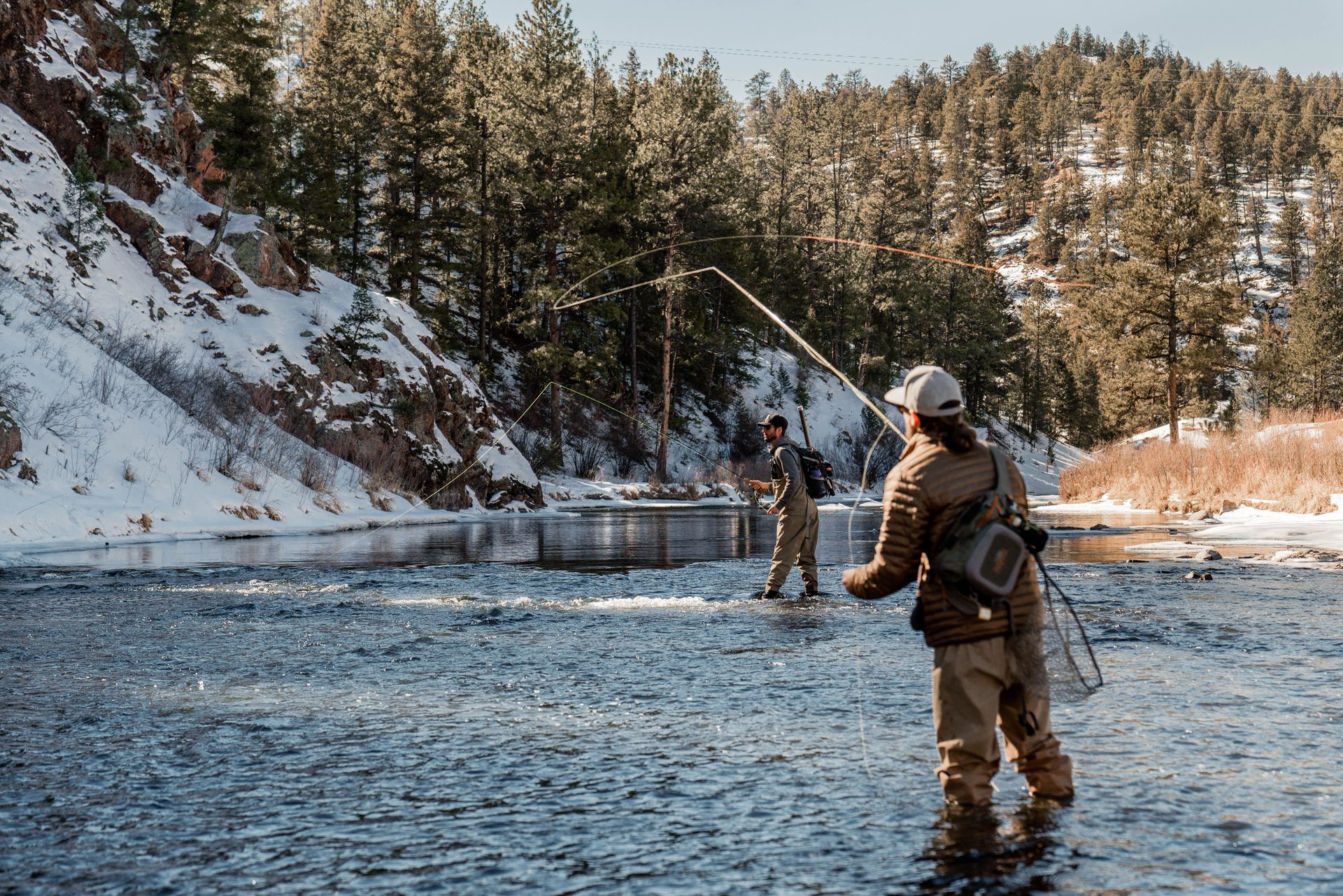 Two people fly fishing in a snowy river, trees and cliffs in the background.