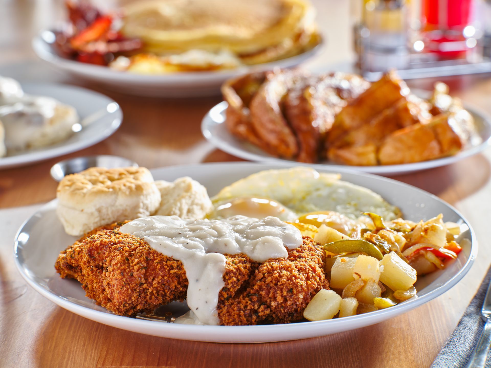 A plate of breakfast with fried chicken, gravy, eggs, potatoes, and a biscuit. Other breakfast dishes in the background.