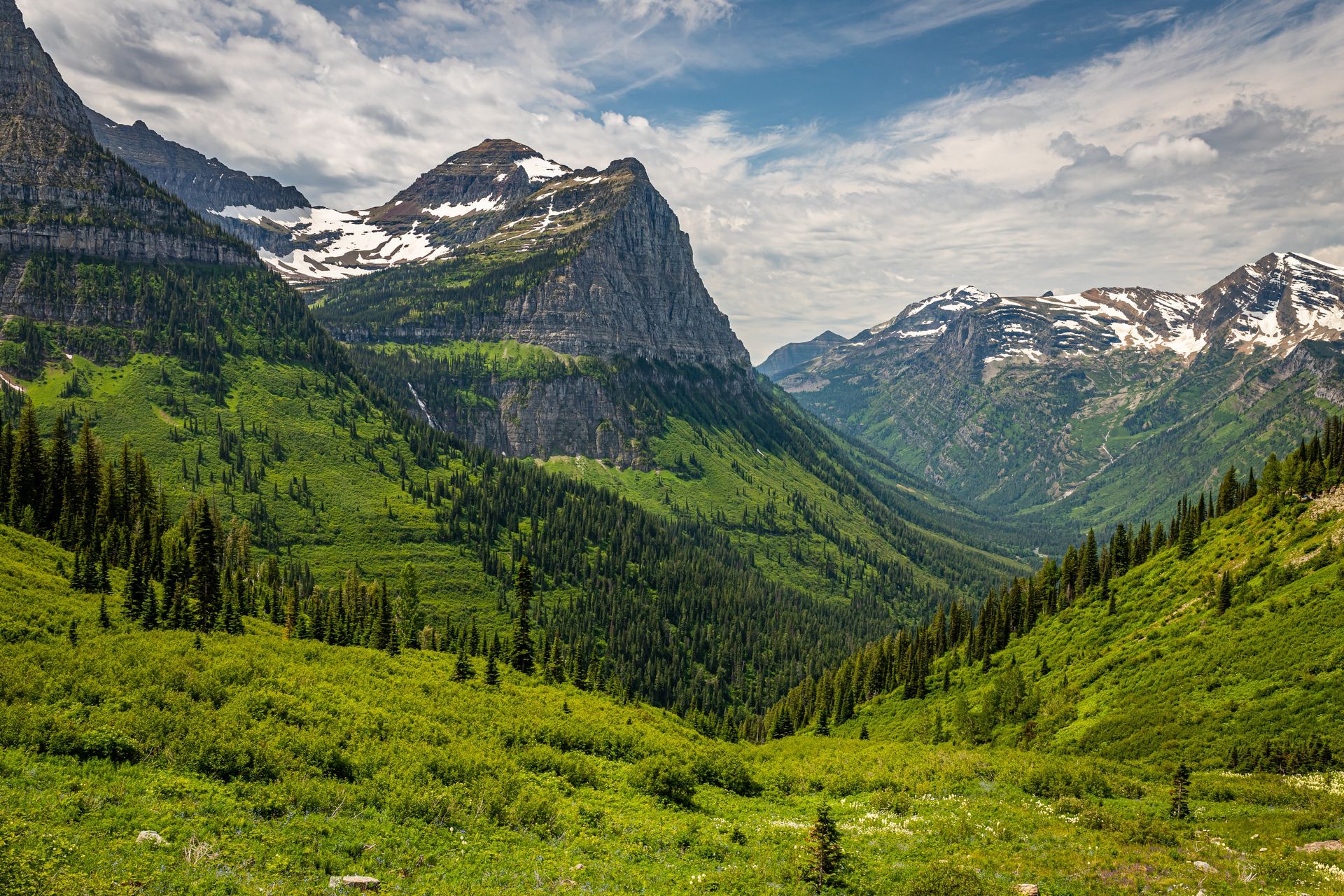 Green valley framed by mountains, trees, and patches of snow under a partly cloudy sky.