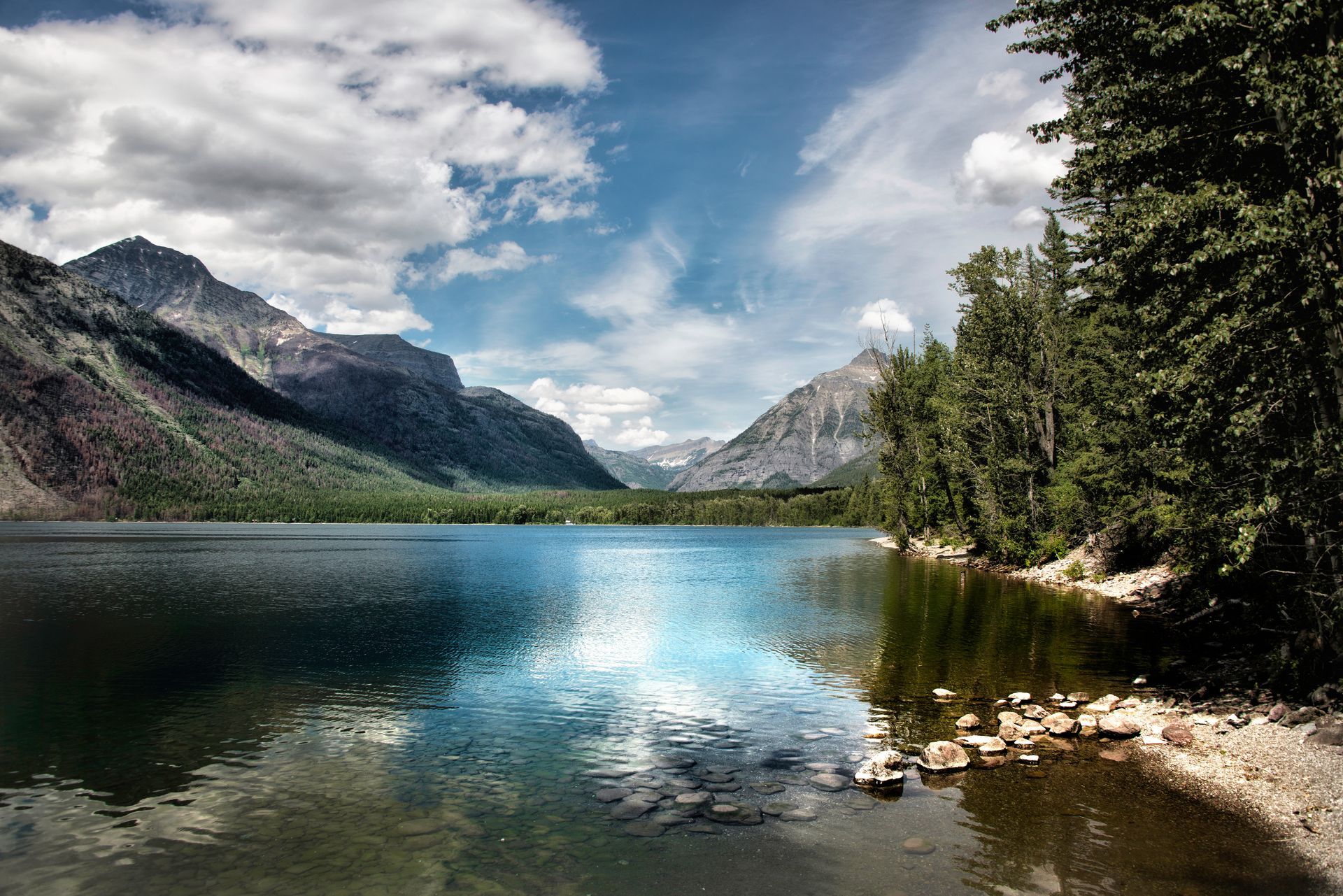 Lake surrounded by mountains and trees, reflecting blue sky and clouds.