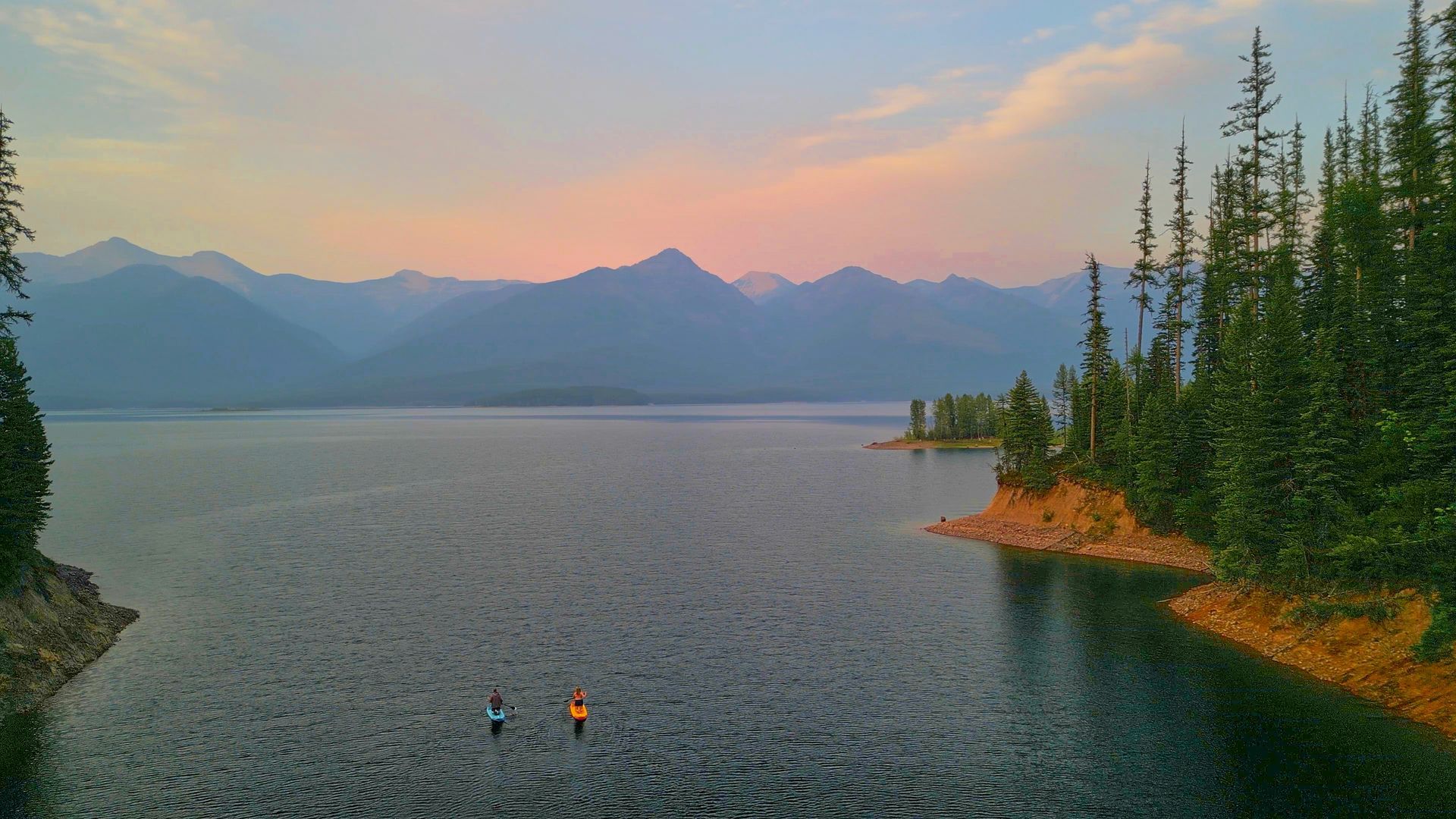 Lake scene at dusk with mountains, evergreen trees, and silhouetted figures on the water.