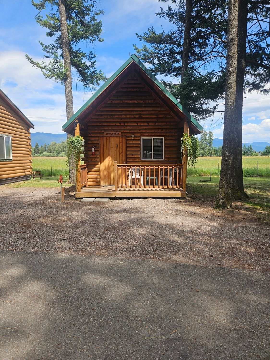 Log cabin with a small porch, set in a rural area with trees and a field; sunny day.