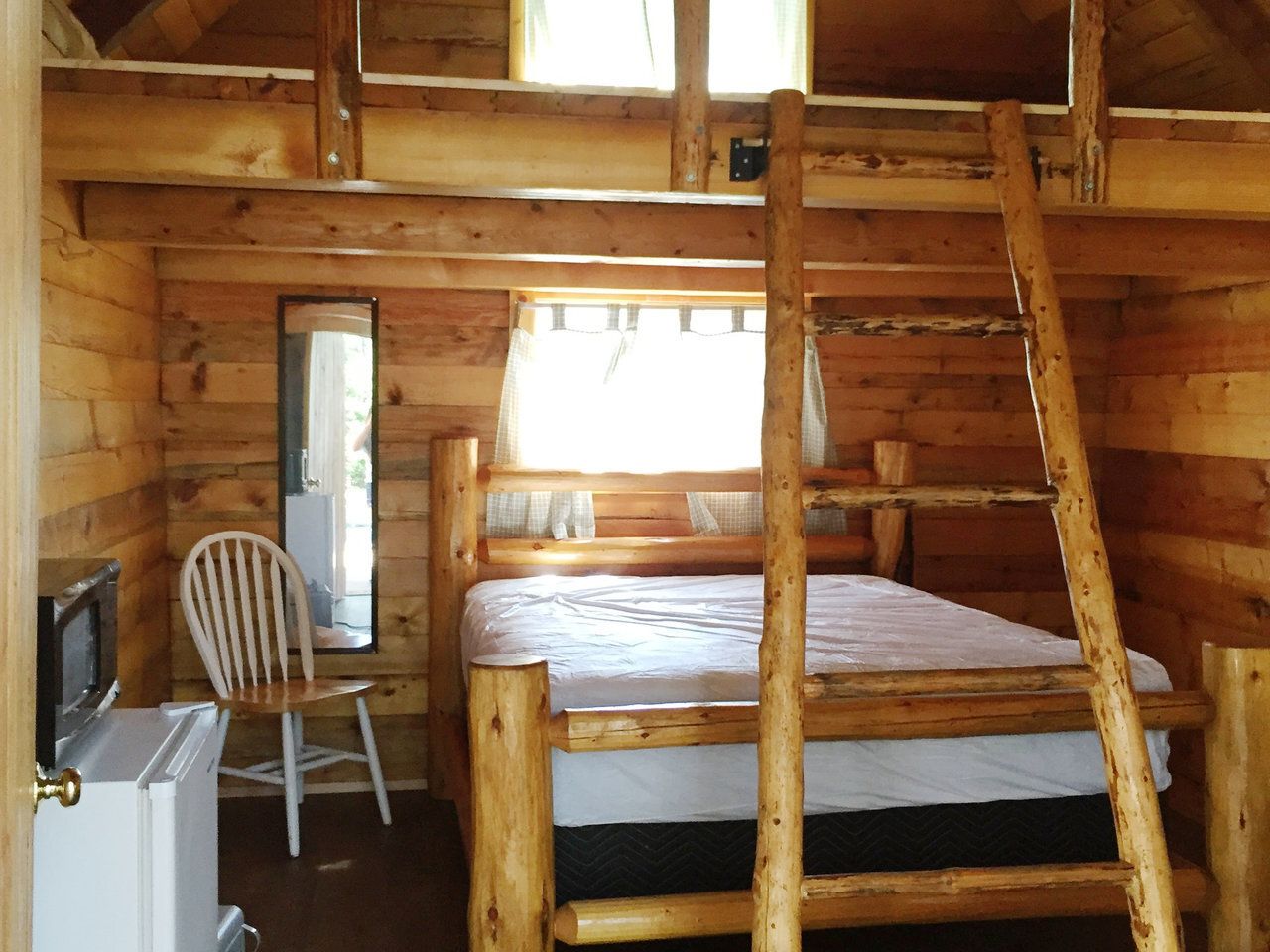 Interior of a wood cabin with a bed, ladder to a loft, chair, mirror, and a small refrigerator.