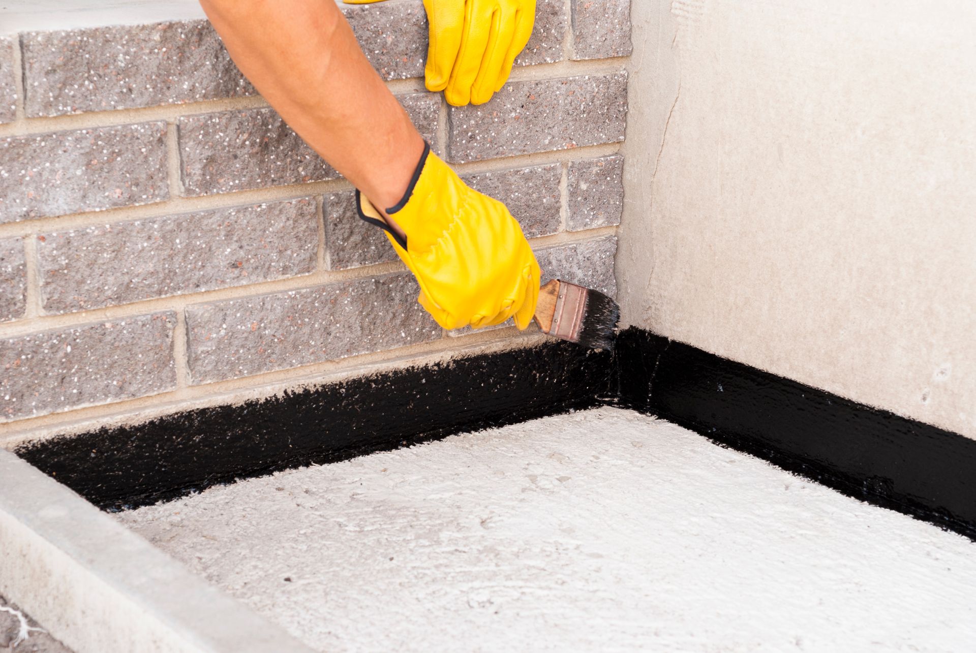 Gloved hands apply black waterproof sealant along brick wall foundation edge.
