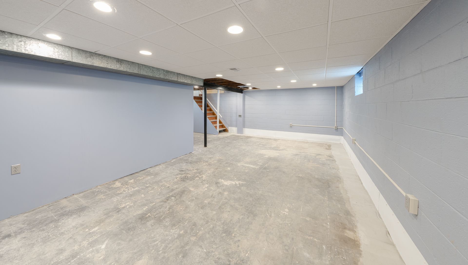 Empty finished basement with concrete floor, recessed ceiling lights, and stairs.