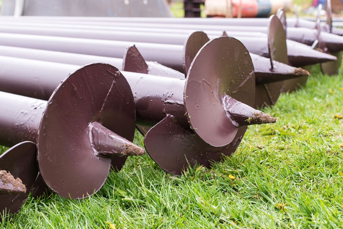 Dark brown helical piers lying on green grass, ready for installation.