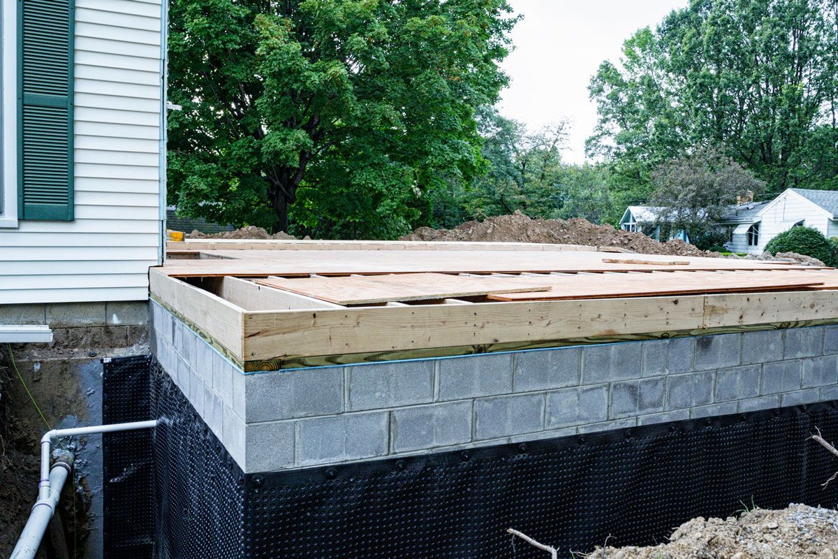 Foundation construction with cinder block walls, wood framing, and black waterproofing membrane. Part of a house is visible on the left.