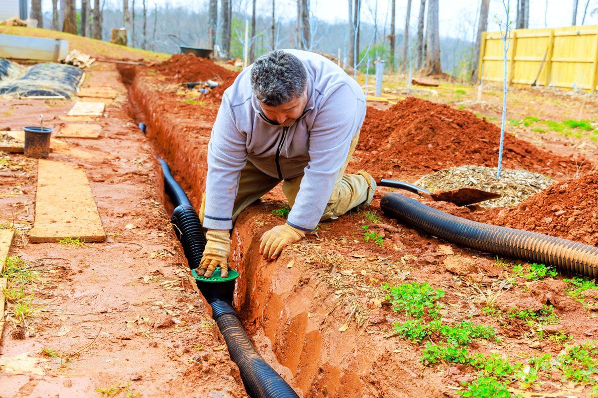 A man kneels in a muddy trench, connecting black drainage pipes. He wears gloves and tan pants in an outdoor setting.