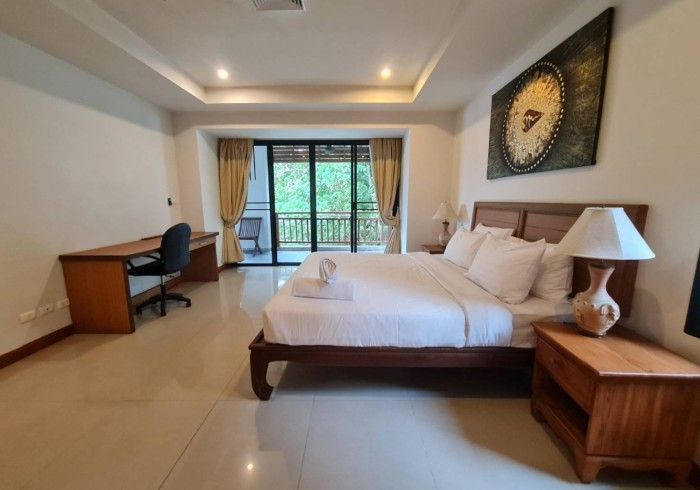 Bedroom with a bed, desk, and balcony access. Wooden furniture, neutral walls, and a decorative tapestry.