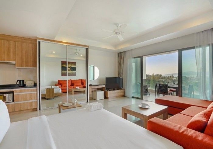 Modern hotel suite with bed, orange couch, balcony, and kitchen area.
