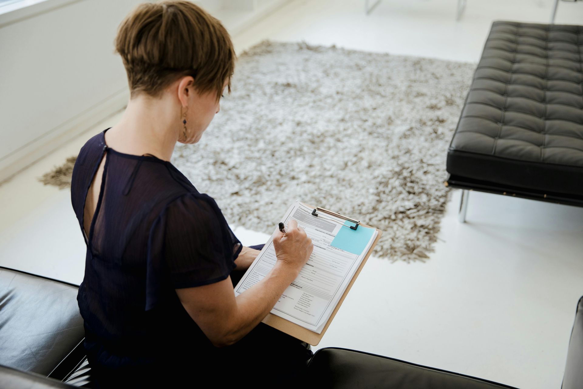 Woman in dark dress writing on clipboard, sitting on black sofa in bright room with rug.