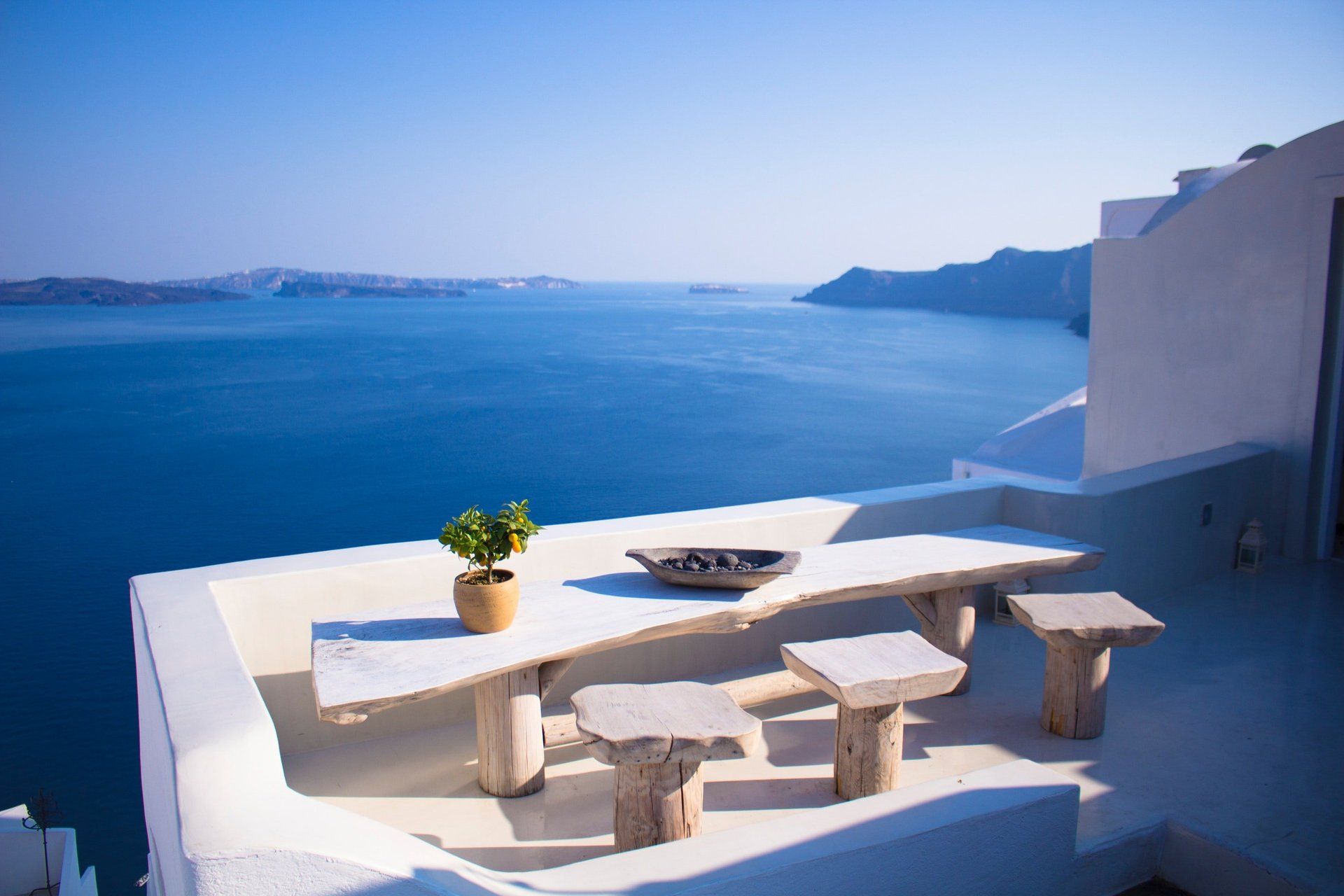 White terrace overlooking blue sea, featuring stone table and stools, potted plant.