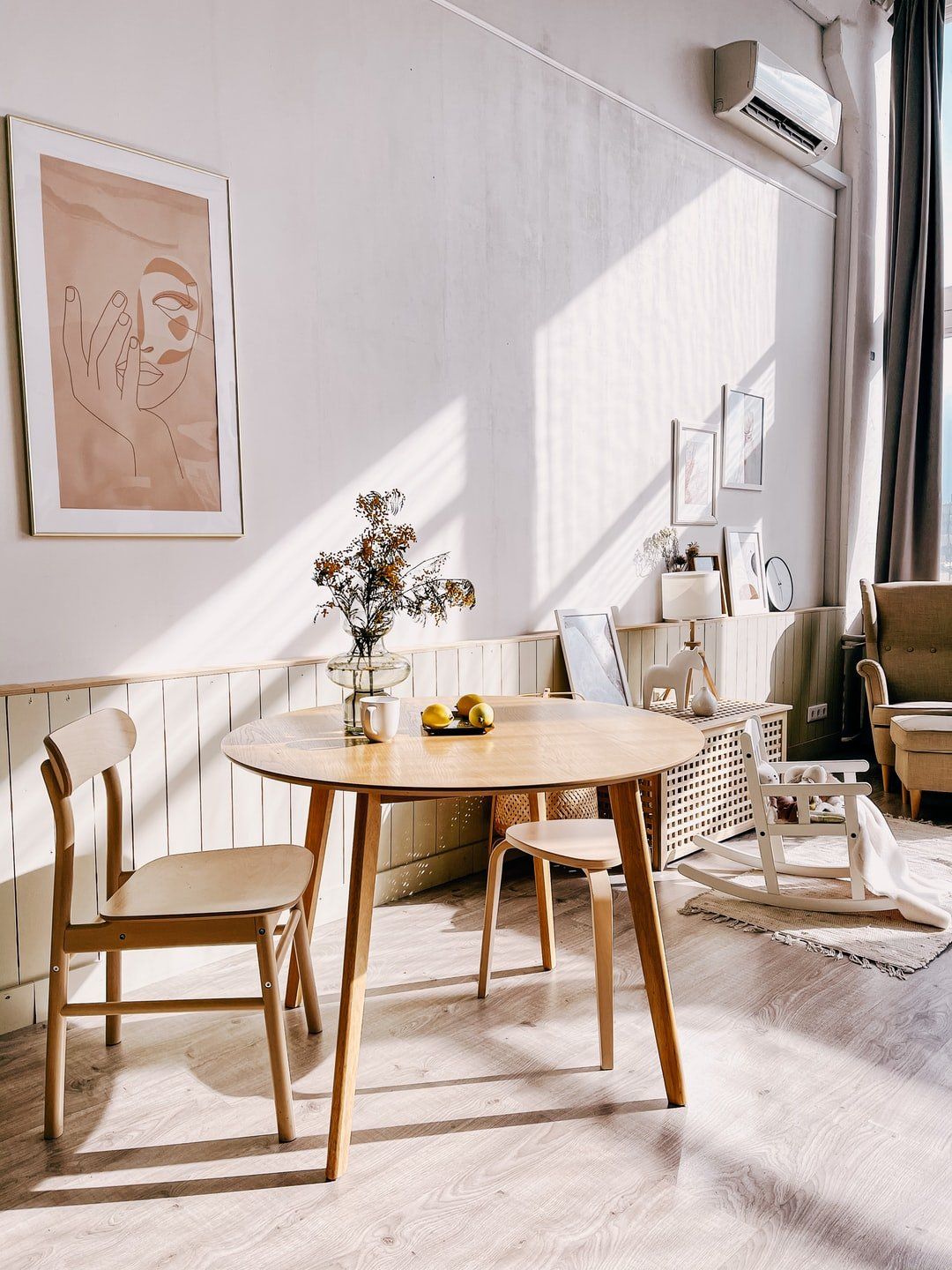 Wooden dining table with chairs, flowers, and artwork in a sunlit room.