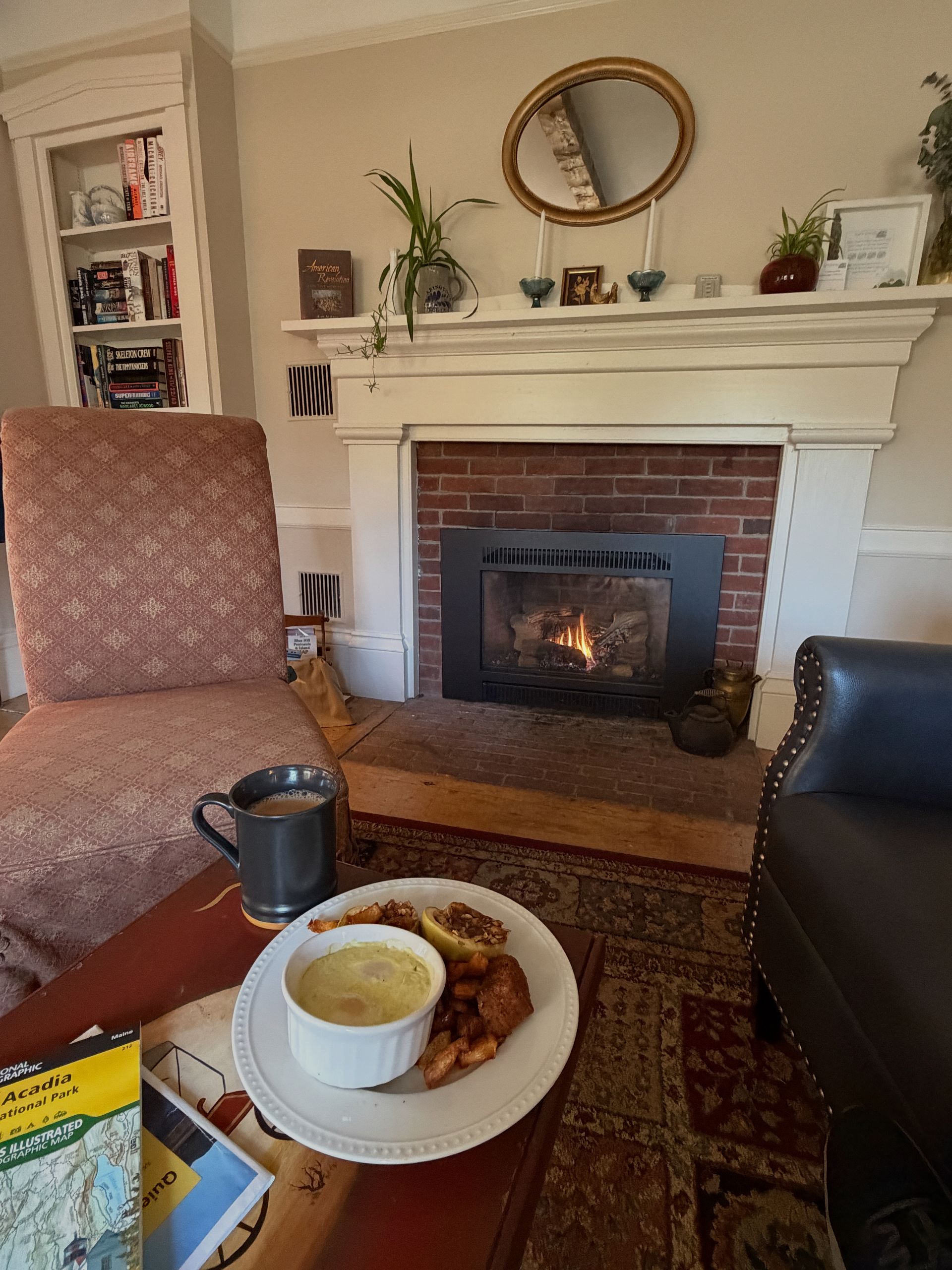 Cozy living room with fireplace, armchair, coffee, and a plate of food. Bookshelf and mirror on the wall.