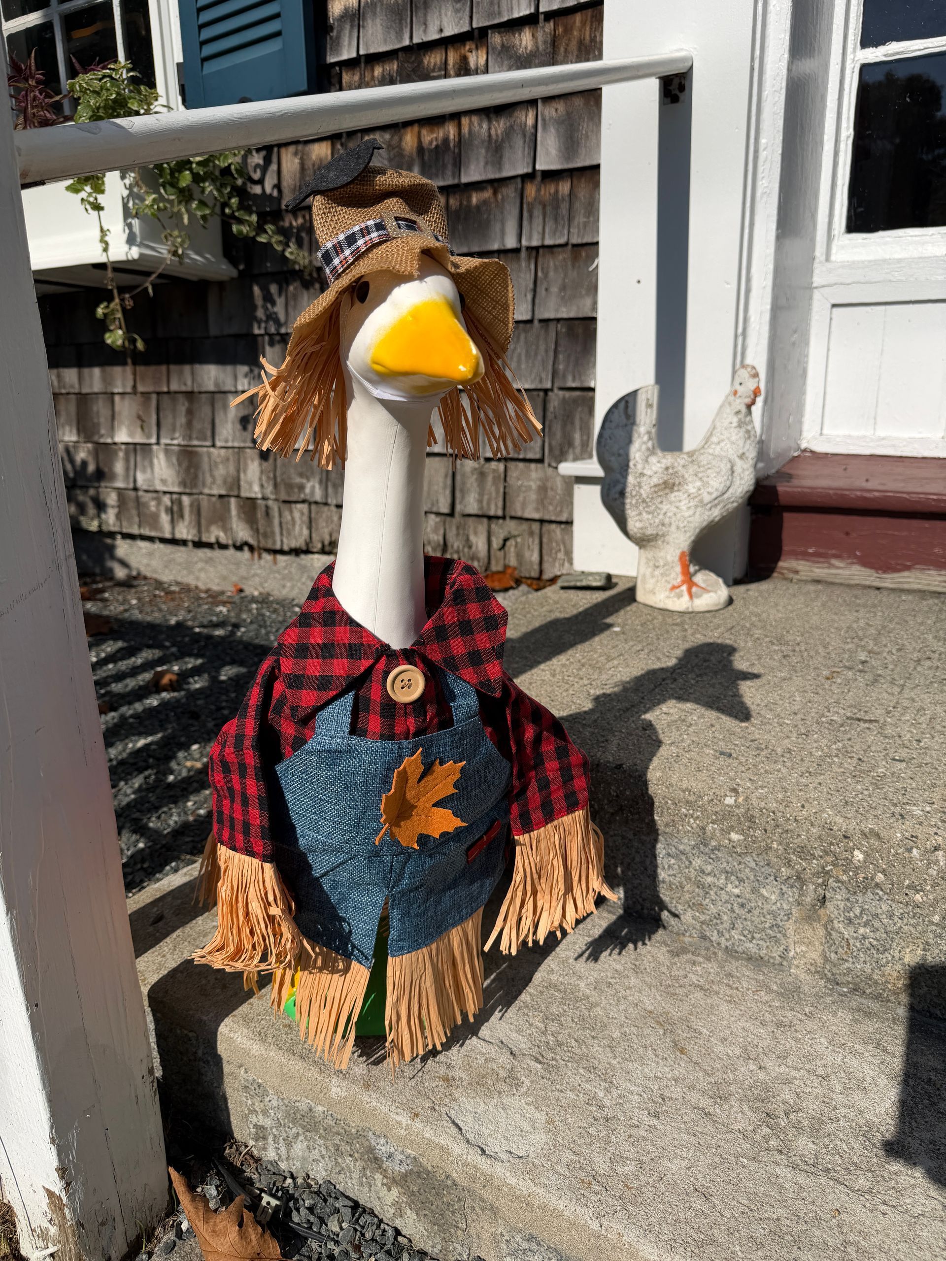 Scarecrow goose statue on porch, wearing overalls, straw hat, and plaid shirt.
