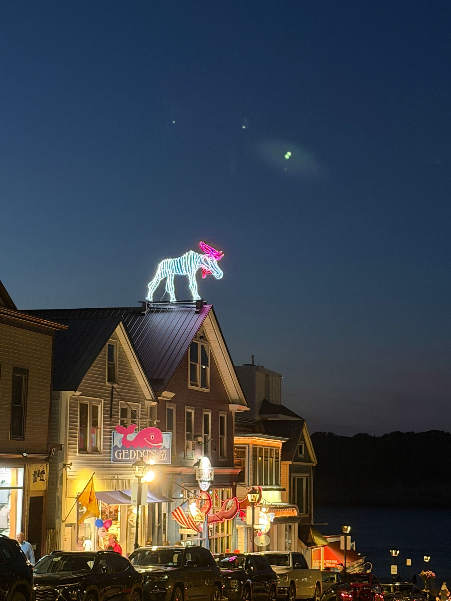 Nighttime street scene with a lit building and a glowing reindeer on the roof under a dark sky.