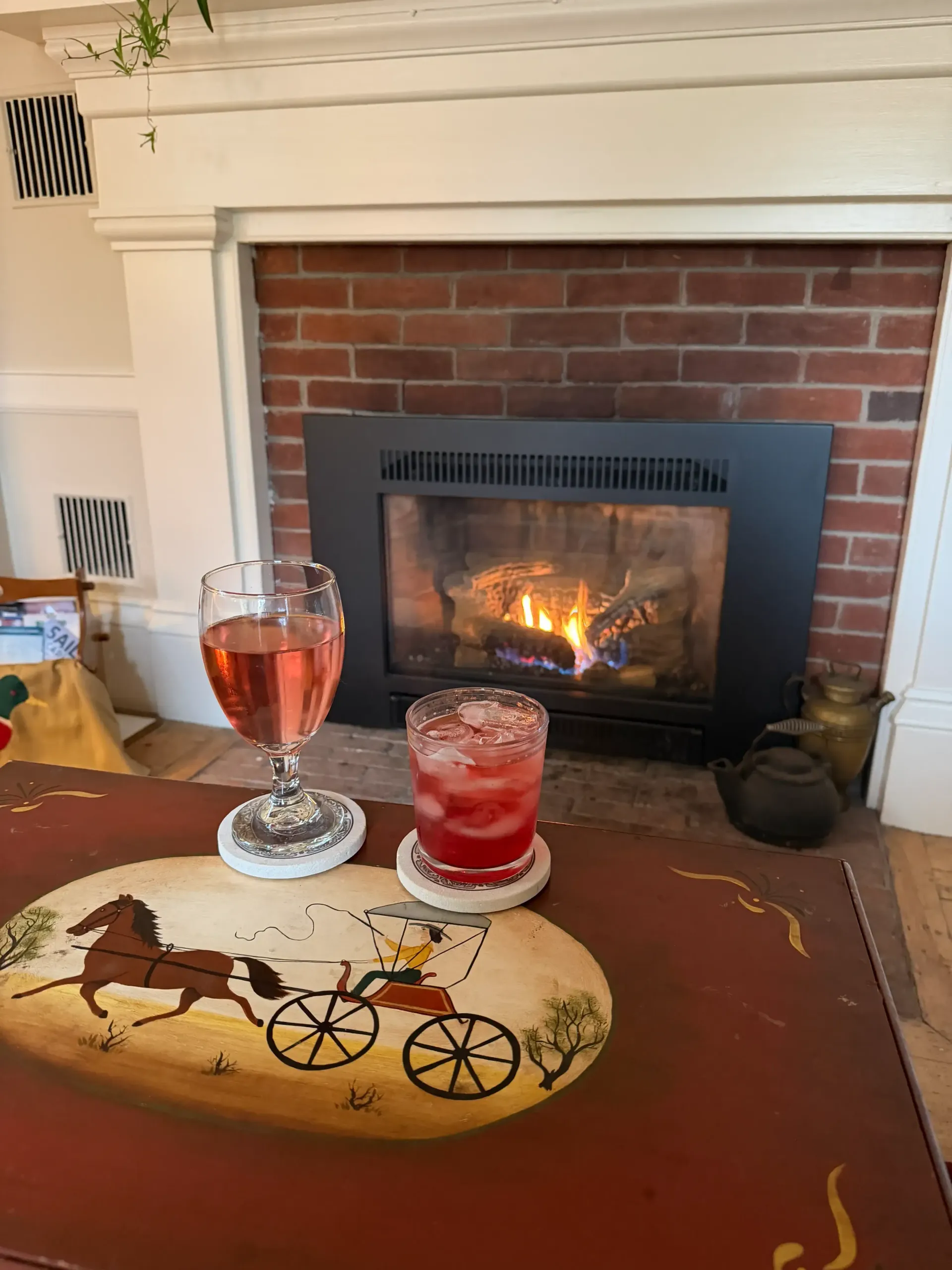 Two cocktails on coasters atop a decorated table, fireplace with burning logs in the background.