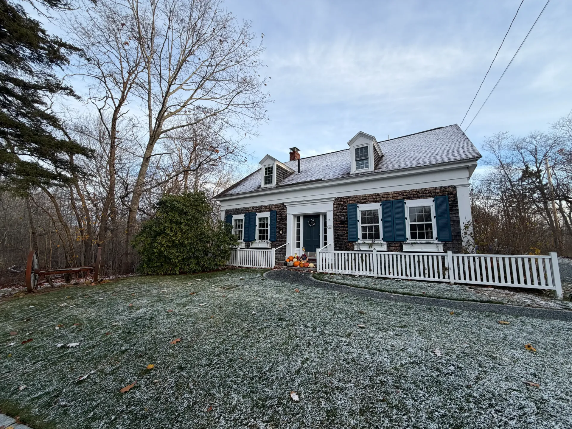 Stone house with blue shutters, a white fence, and snow-covered lawn on a cloudy day.