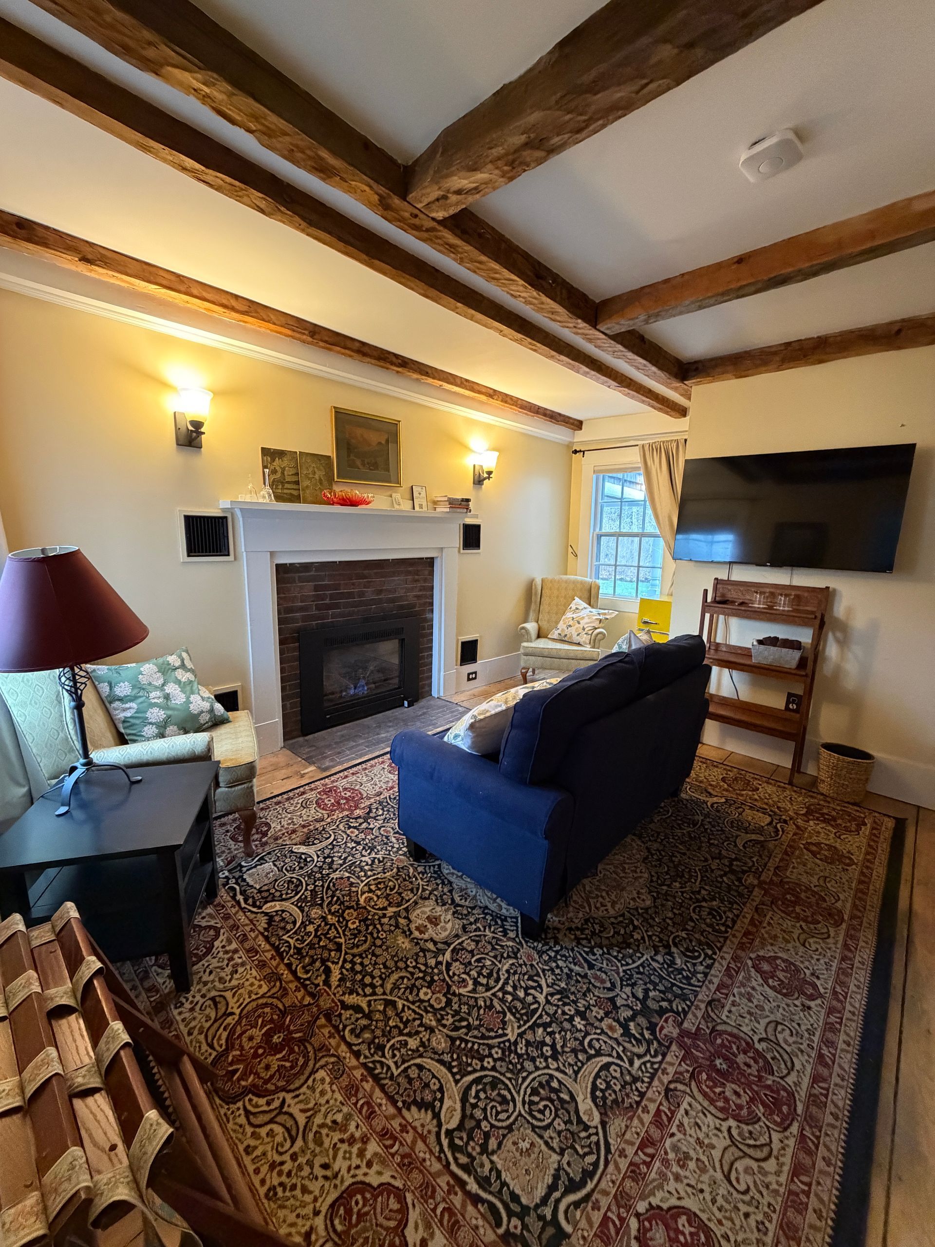 Cozy living room with a fireplace, blue sofa, patterned rug, wall TV, and exposed wood beams.