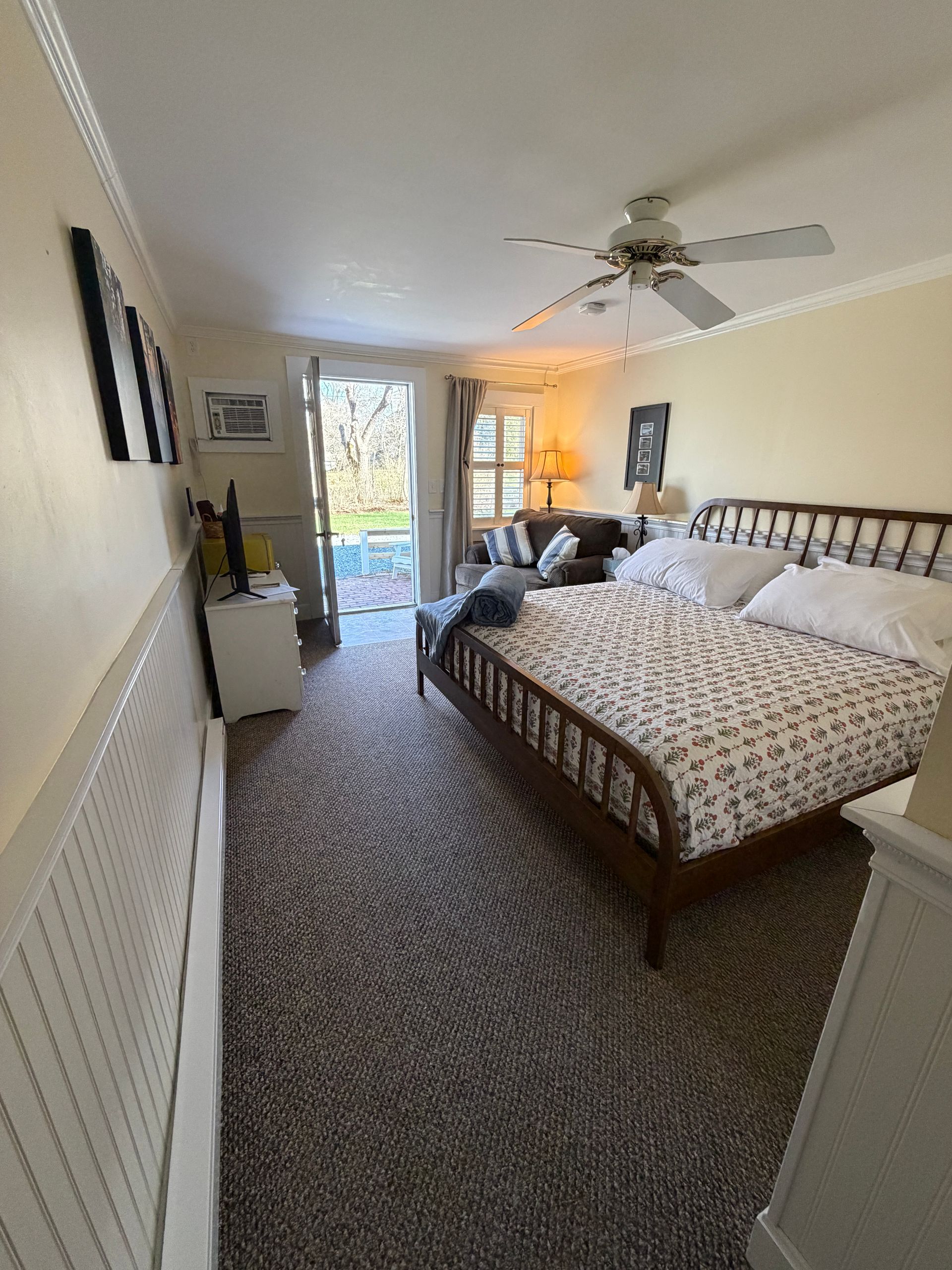 Bright bedroom with a king bed, ceiling fan, carpeted floor, and open door to a sunny patio.