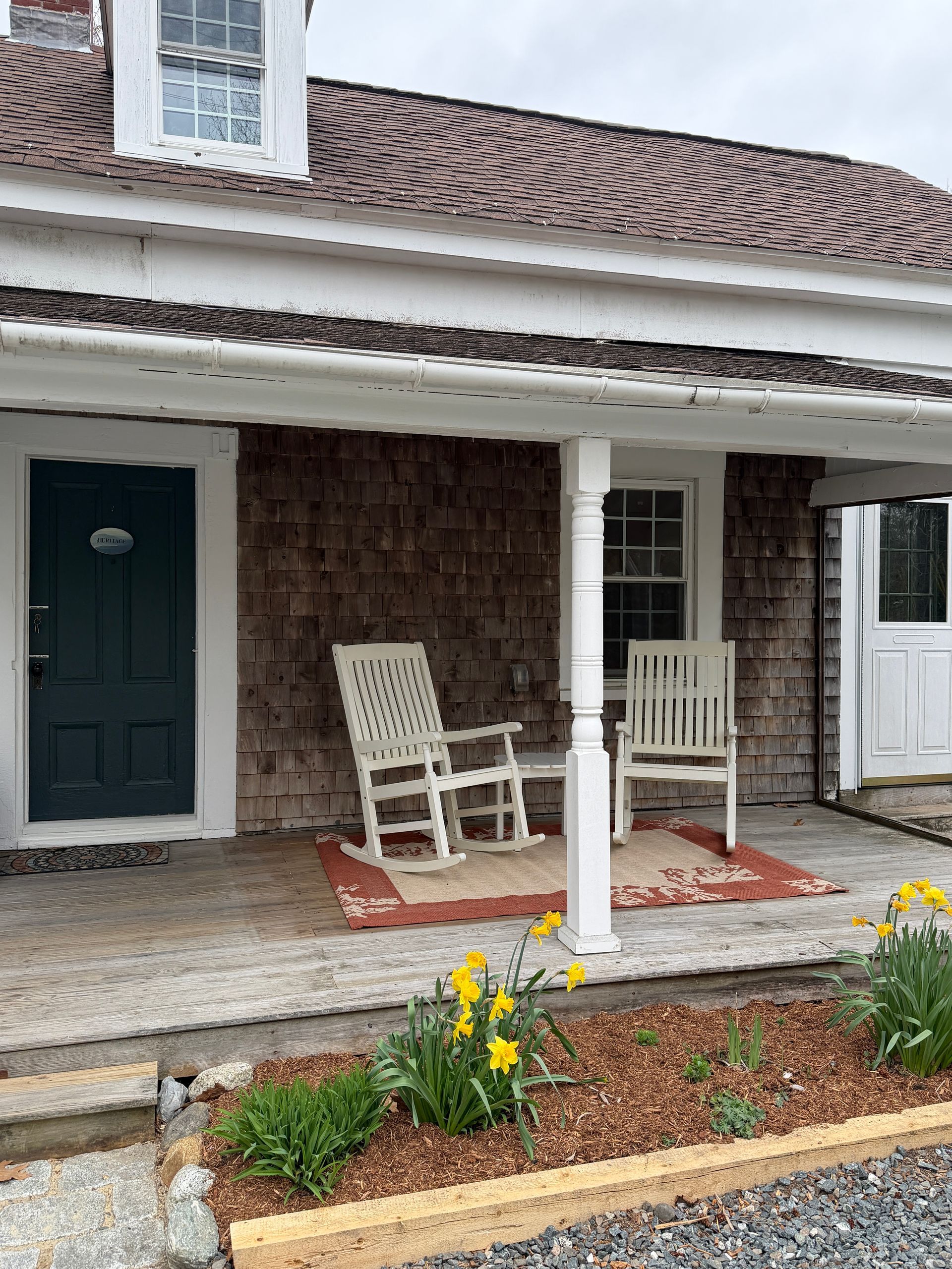 Porch with two white rocking chairs, dark green door, and yellow flowers in a landscaped bed